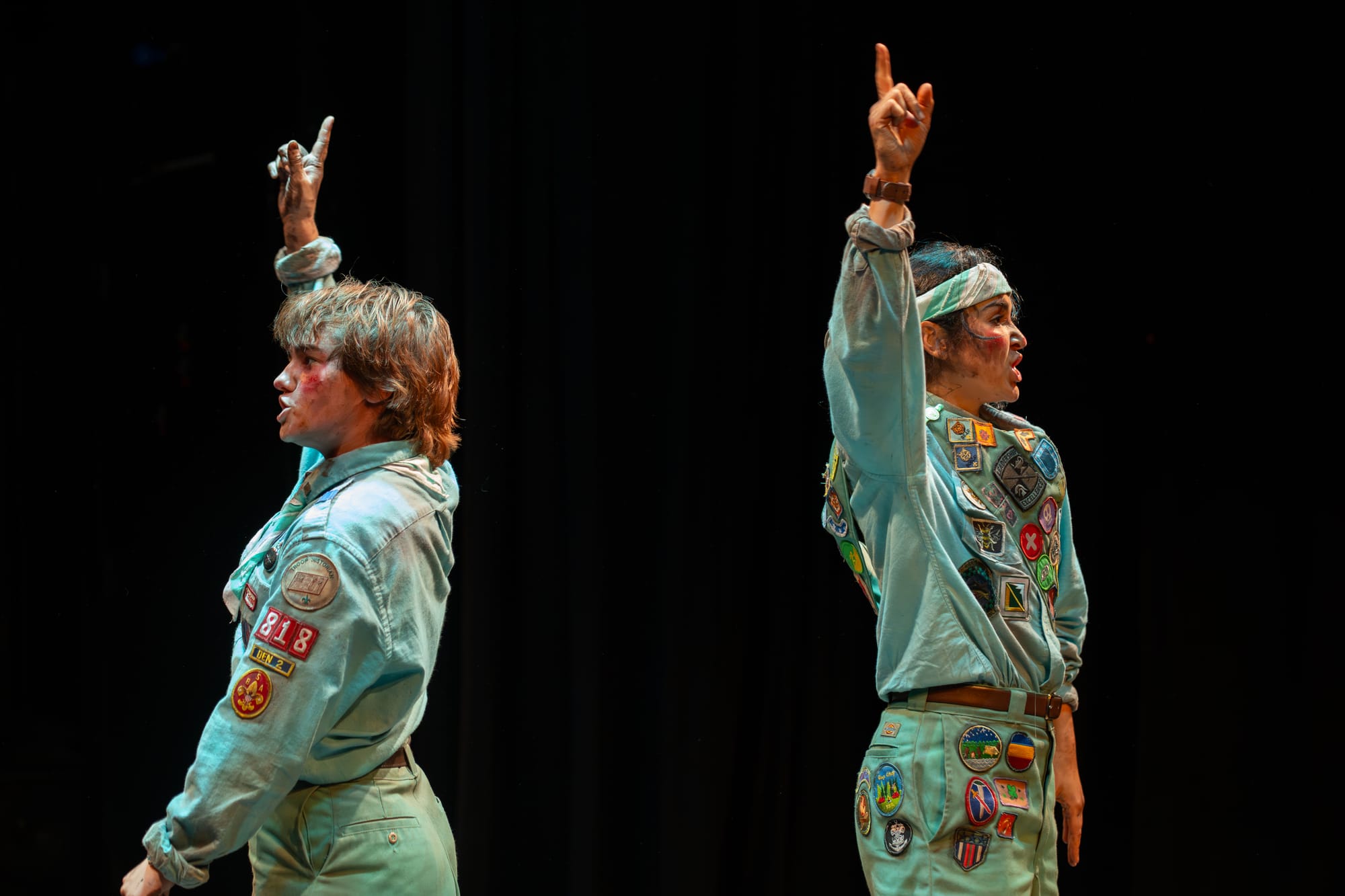 Two performers stand on a dark stage wearing green scout uniforms. They both have one arm pointed upwards.