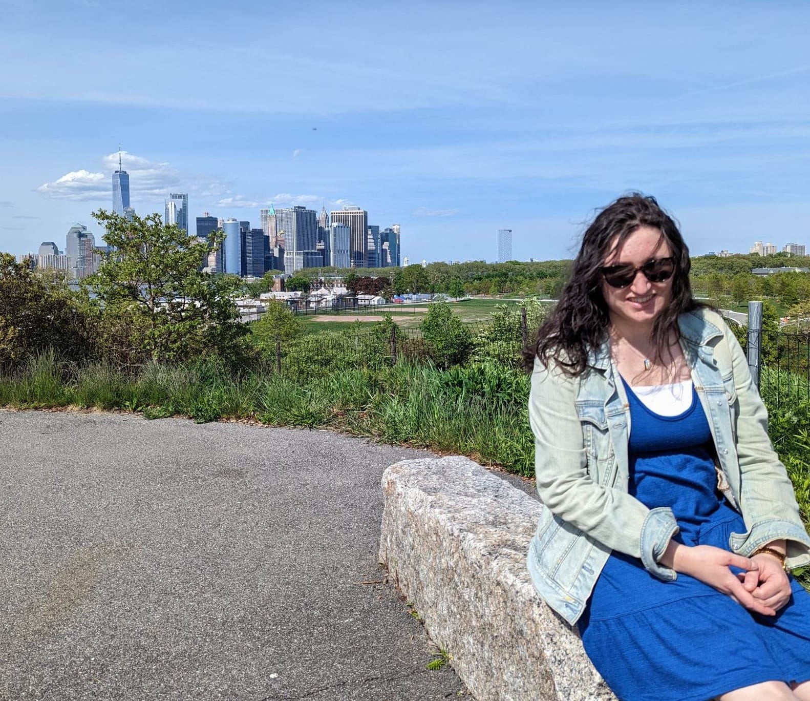 A pale Caucasian woman with wavy brown hair sits on a rock and smiles at the camera. Sunglasses are covering her eyes. She is wearing a blue dress and a denim jacket. The Manhattan skyline is in the background.