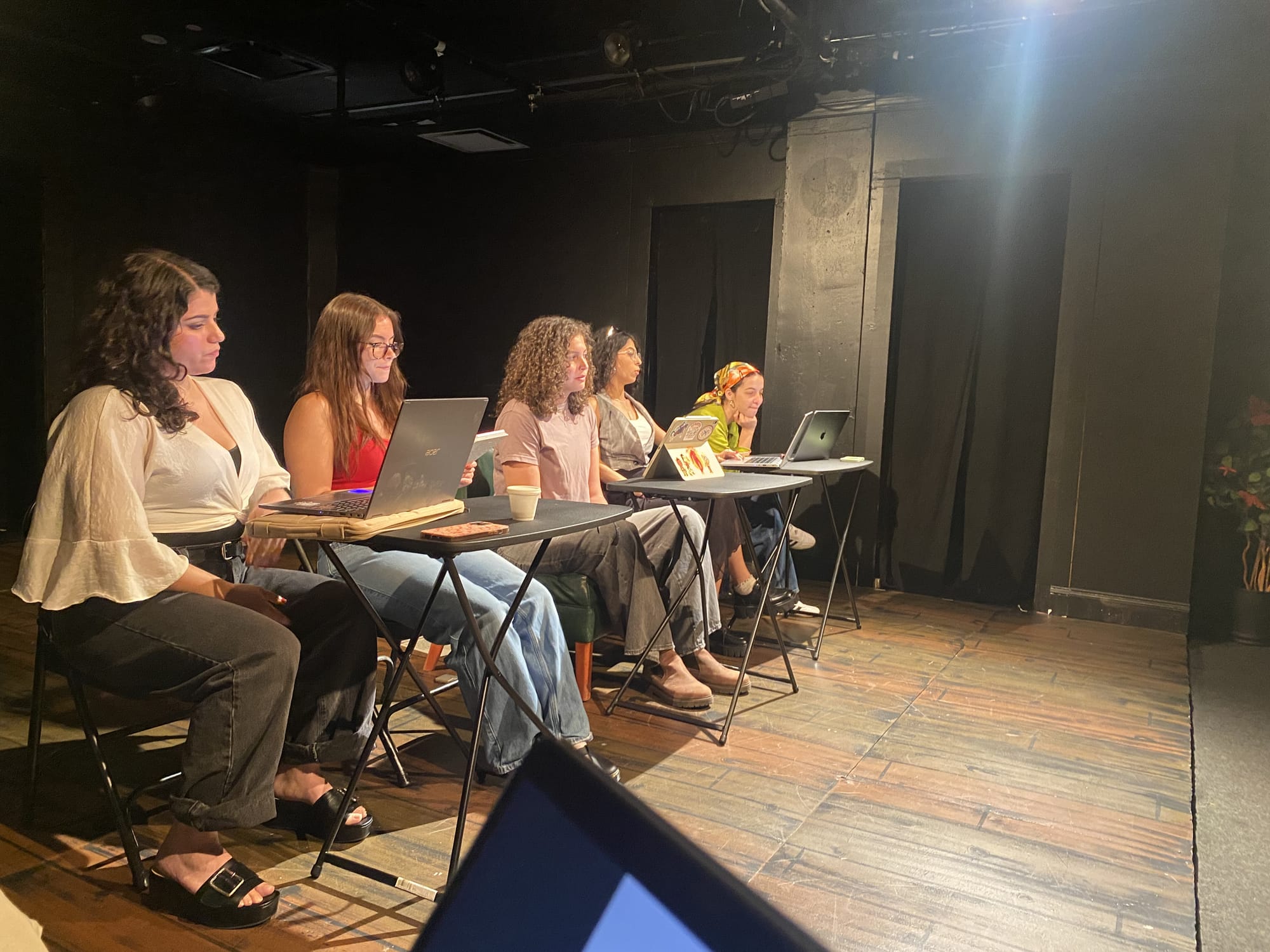Five women-presenting actors sit at tables in a theatre for a staged reading.