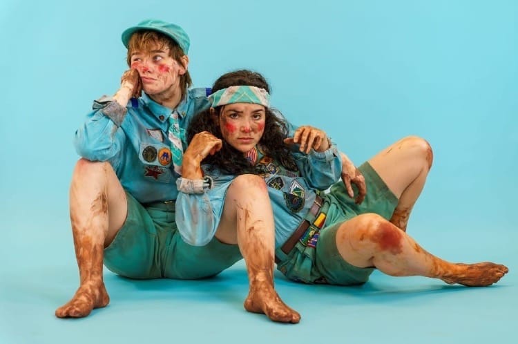 Two performers in scout uniforms covered in mud sit on the floor.